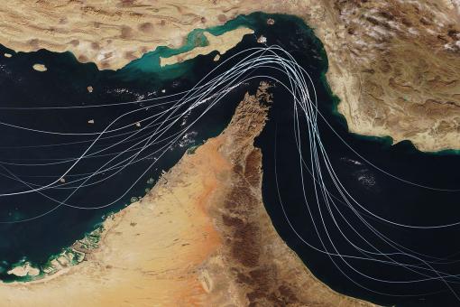 Satellite view of the Strait of Hormuz with white graphic lines representing global shipping lanes and maritime traffic between the Persian Gulf and Gulf of Oman.