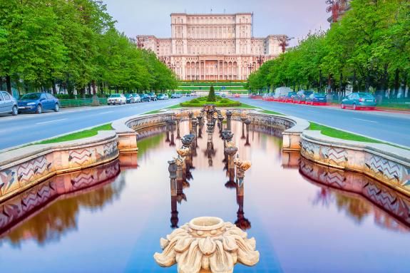 Photograph of fountains along the tree-lined Bulevardul Unirii, a major thoroughfare in central Bucharest, with the Palace of the Parliament in the background at dawn.