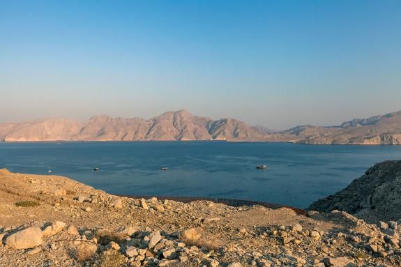 View across the Strait of Hormuz toward rugged mountains of the Musandam Peninsula in Oman. Small boats rest on the calm water beneath the arid landscape of the northern Arabian Peninsula.